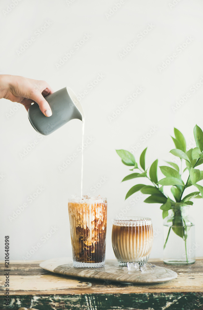 Iced coffee in tall glasses with milk poured over from pitcher by hand, white wall and green plant b