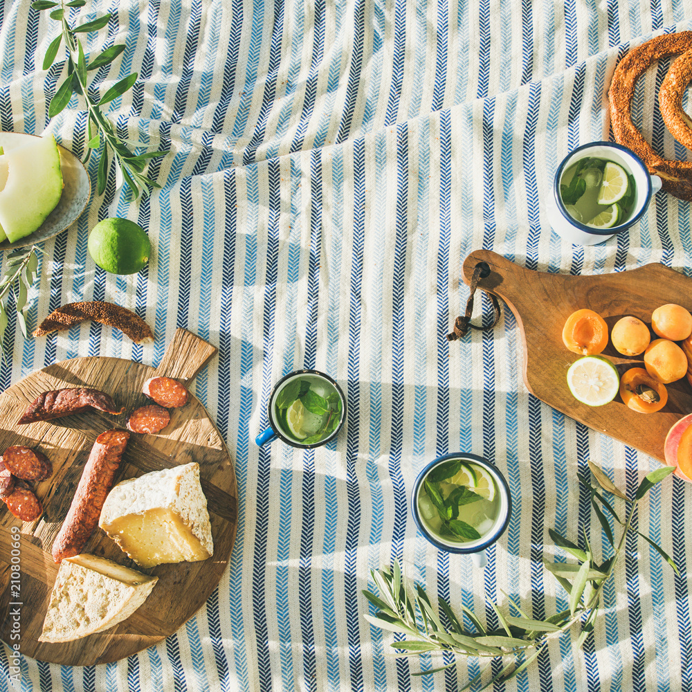 Flat-lay of summer picnic set with fruit, cheese, sausage, bagels and lemonade over striped blanket,