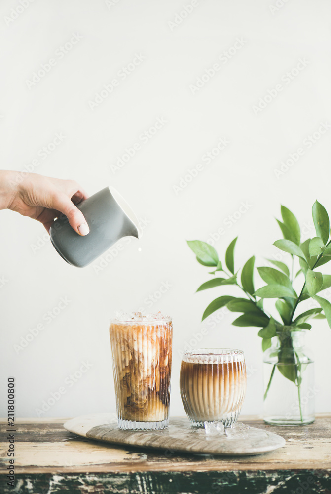 高玻璃杯中的冰咖啡，用手从水罐中倒出牛奶，白墙和绿色植物膜