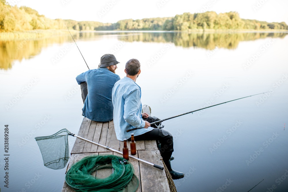 两个穿着蓝色衬衫的男性朋友坐在木码头上，用网和鱼竿一起钓鱼