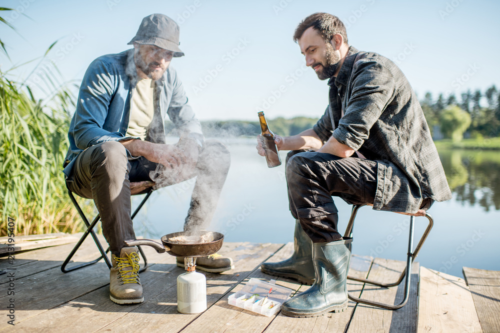 两名渔民在湖附近的木码头上野餐时，坐着用啤酒煎鱼