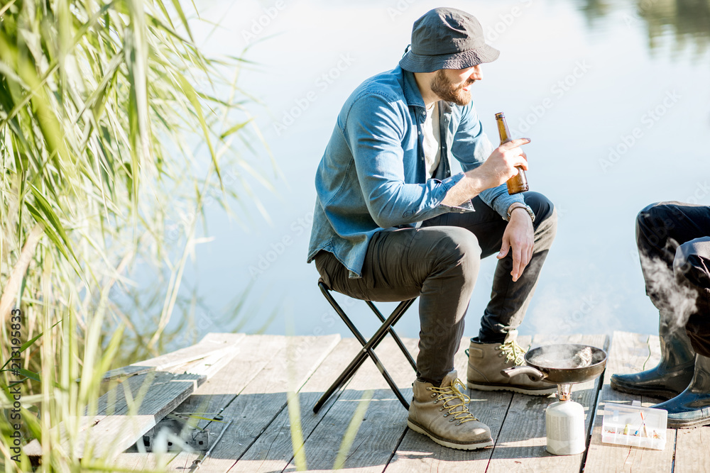 Two fishermen frying fish sitting with beer during the picnic on the wooden pier near the lake in th