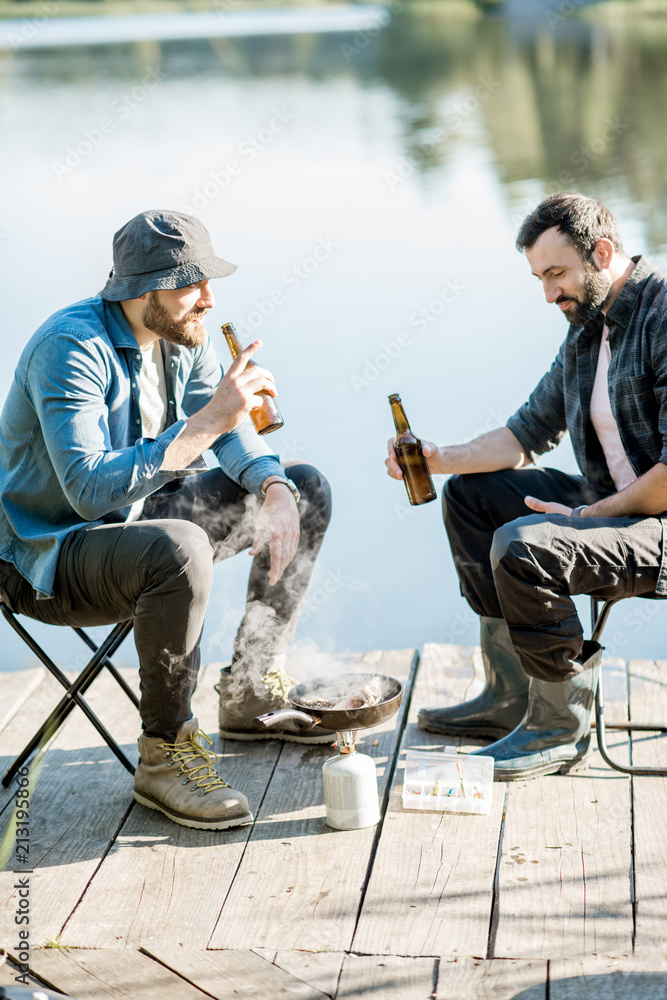 两名渔民在湖附近的木码头上野餐时坐着用啤酒煎鱼