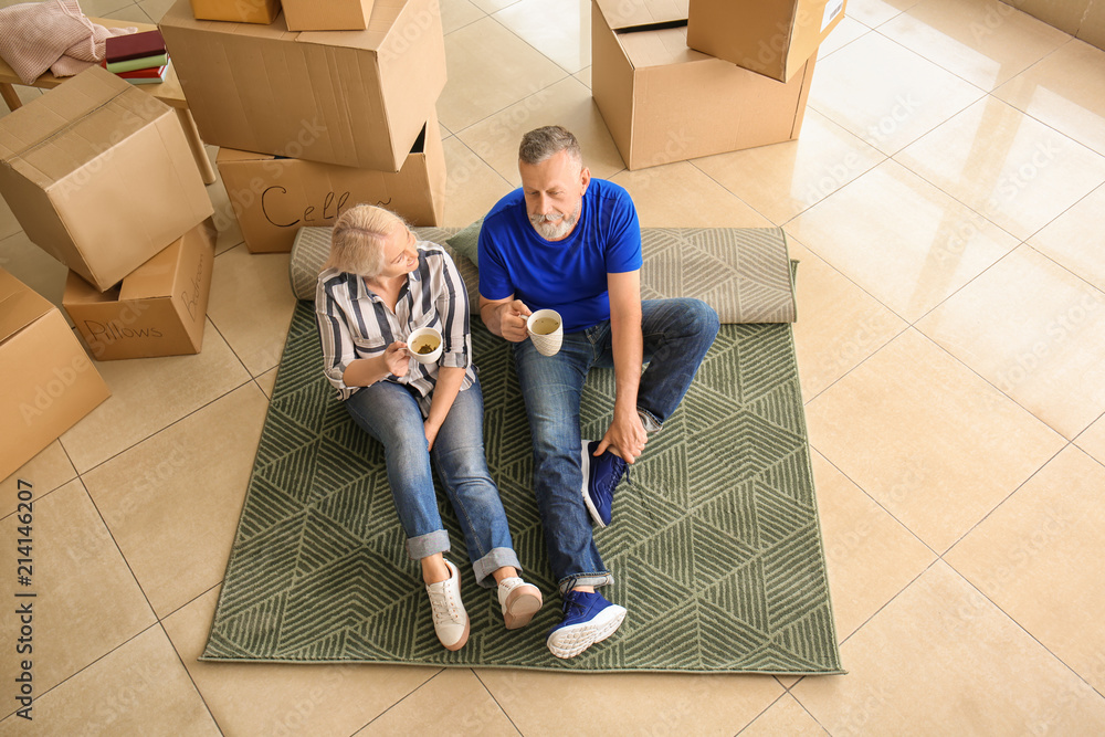 一对带着物品的成熟夫妇坐在地毯上，在室内喝茶。搬进新房子