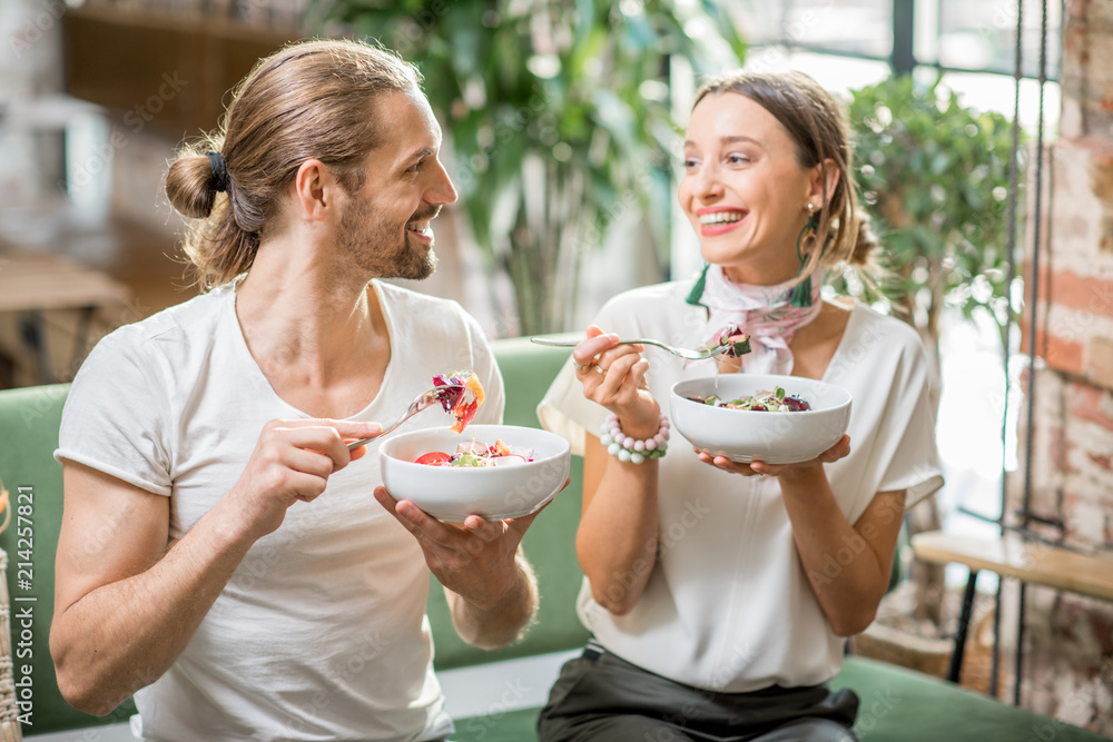 幸福的素食夫妇穿着白色t恤，坐在室内的草地上吃着健康的沙拉