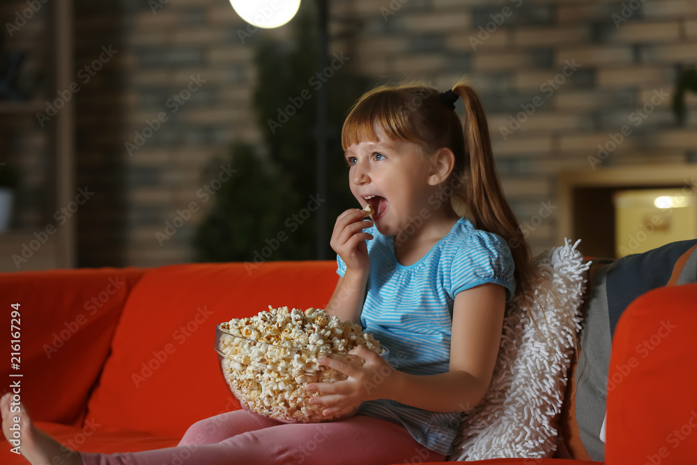 小女孩晚上在沙发上看电视时吃爆米花