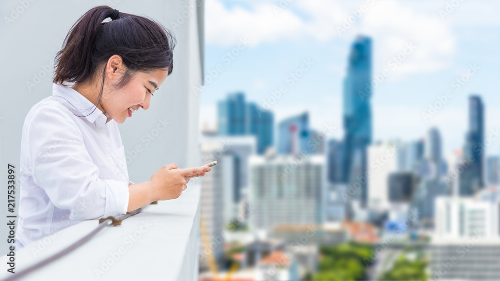 快乐的年轻亚洲女人站在屋顶上用手机，模糊的繁忙城市背景