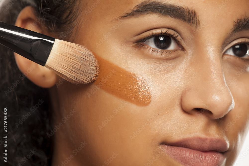 一位年轻的深色皮肤女性在白色皮肤上用刷子在脸上涂液体化妆品的肖像