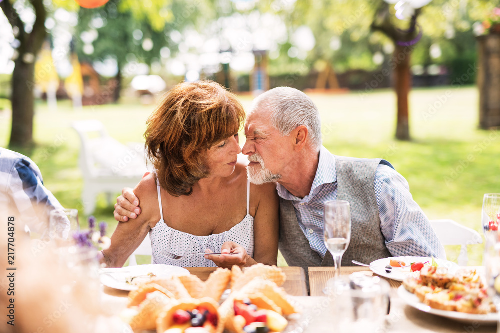 一对老年夫妇在花园聚会上坐在桌子旁，抚摸着鼻子。
