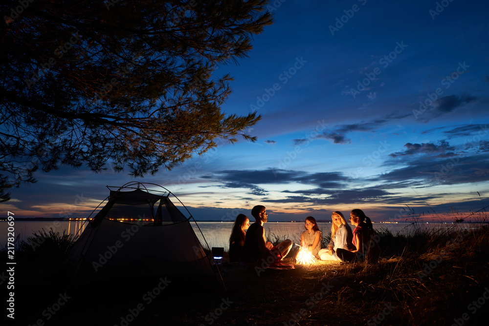 湖岸夏季夜间露营。五名年轻游客一组坐在篝火旁的海滩上