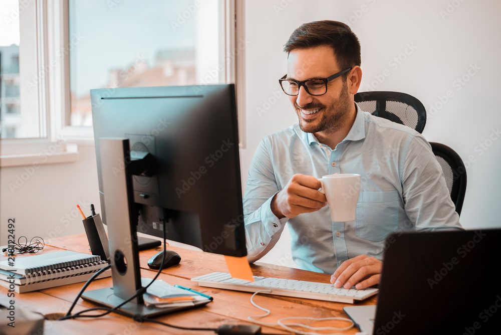 英俊的男人一边喝咖啡，一边看着家里的电脑显示器。