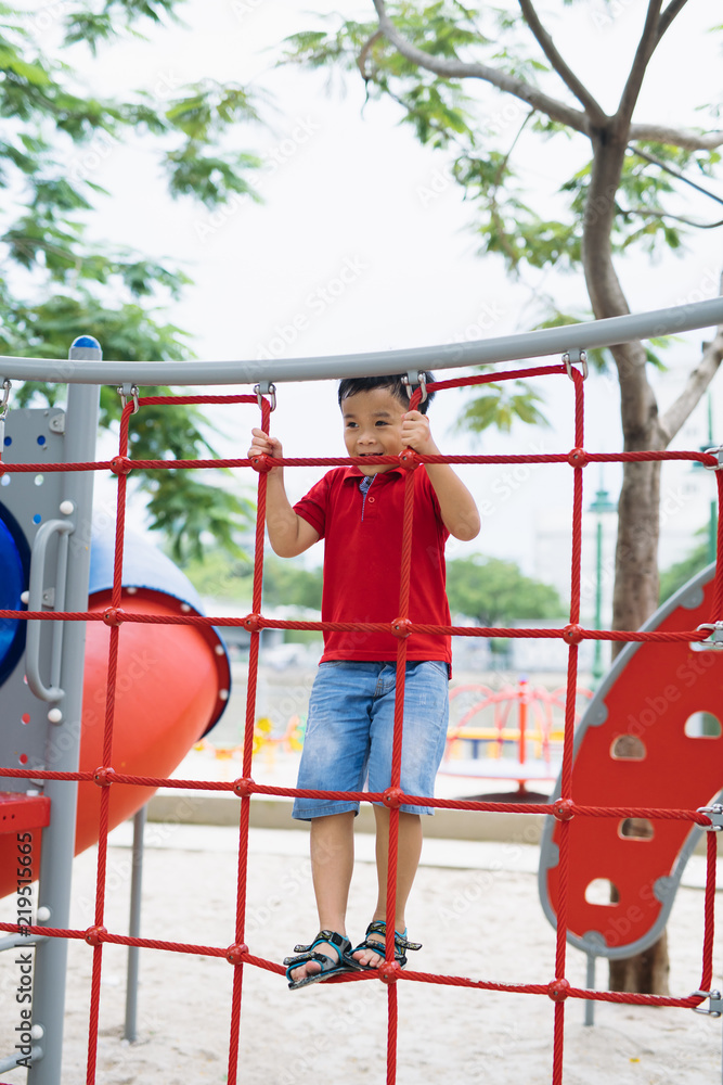 年轻的亚洲男孩用手爬上红绳围栏和灰色横杆，在户外玩耍时锻炼