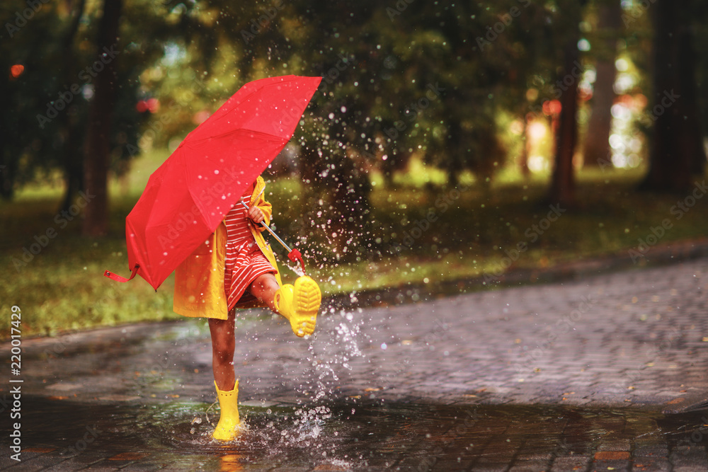 在秋天散步时，快乐的女孩在水坑里撑着伞和橡胶靴