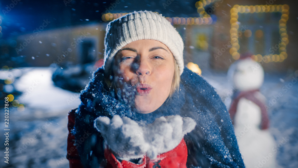快乐的冬夜，大笑的年轻女子从手中吹雪。背景是Idylic Hous