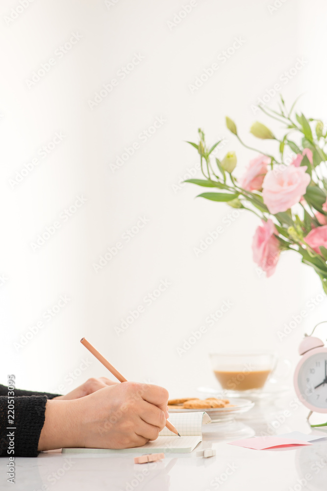 女性在桌子旁的笔记本上写字，俯视图。白色桌子上摆放着鲜花