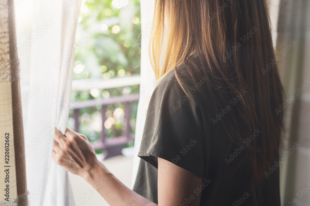 年轻的亚洲女人在早晨揭开窗帘，看着窗外