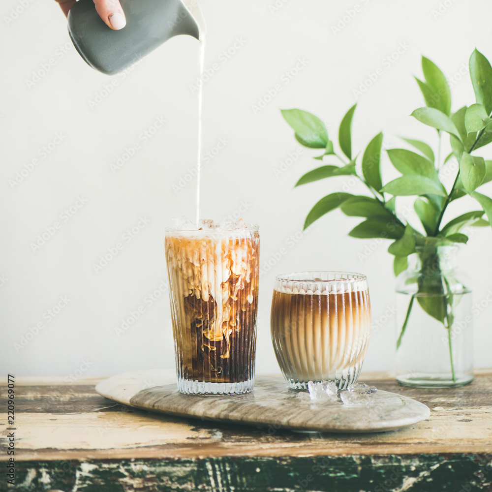 高玻璃杯里的冰咖啡，用手从水罐里倒出来的牛奶，白墙和绿色植物b