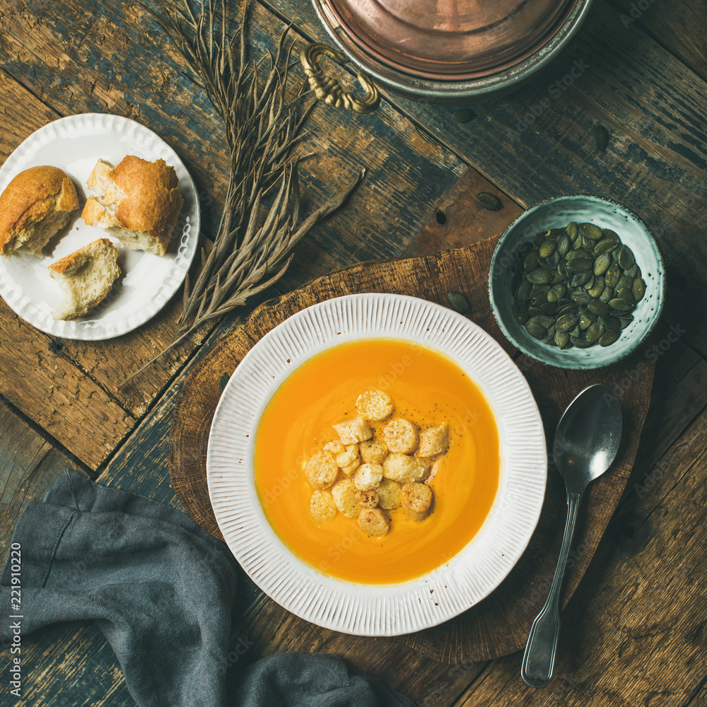 Flat-lay of fall warming pumpkin cream soup with croutons and seeds on board over rustic wooden back