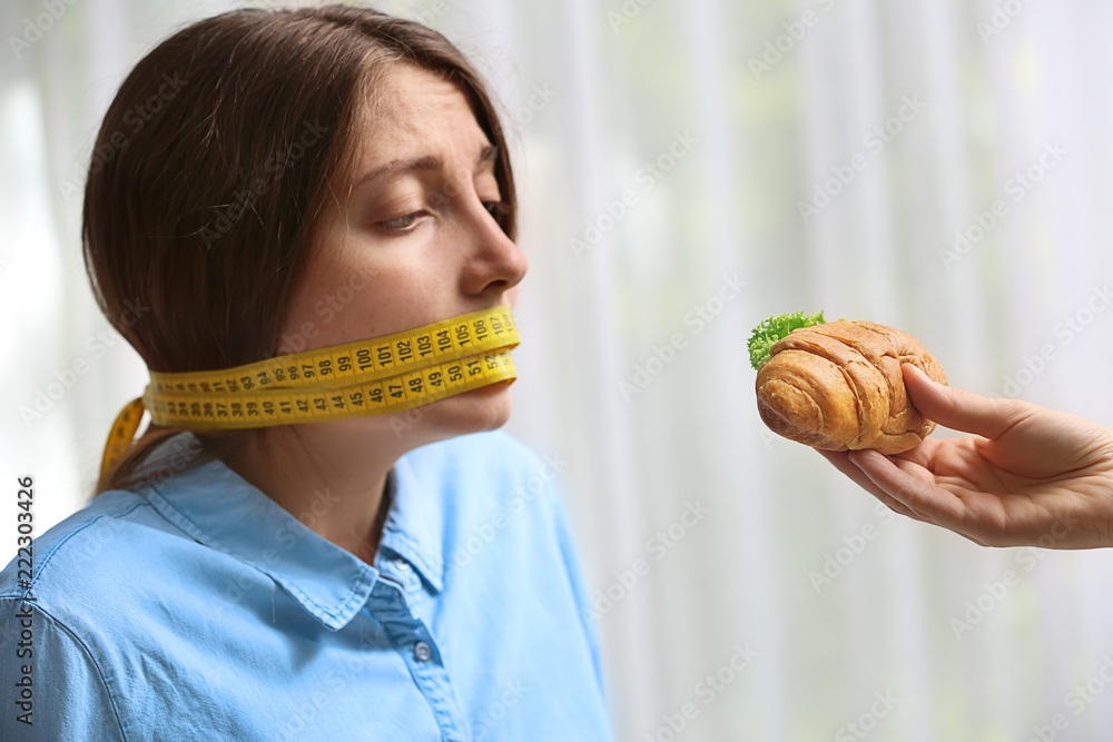 Offering of tasty croissant sandwich to young woman with measuring tape around her mouth on light ba