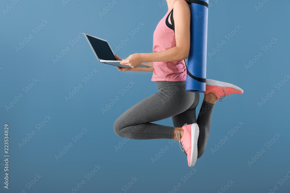 年轻女子拿着瑜伽垫和笔记本电脑在彩色背景上跳跃。休息和休息之间的平衡概念