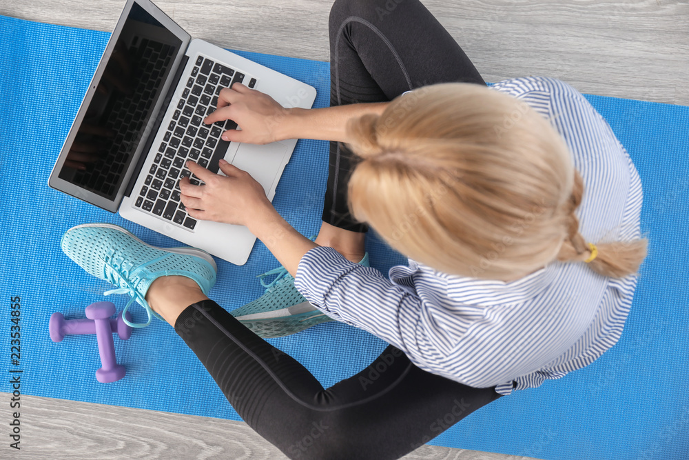 年轻女子拿着笔记本电脑坐在室内瑜伽垫上。休息和工作之间的平衡概念