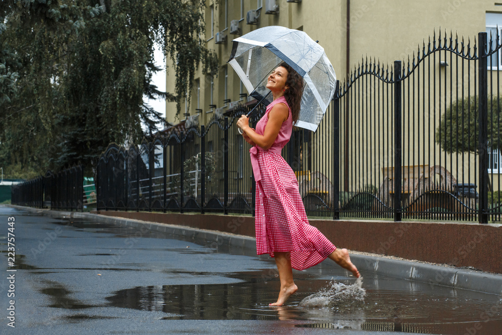 年轻女孩穿着红色连衣裙，打着透明雨伞，站在水坑里雨中跳舞