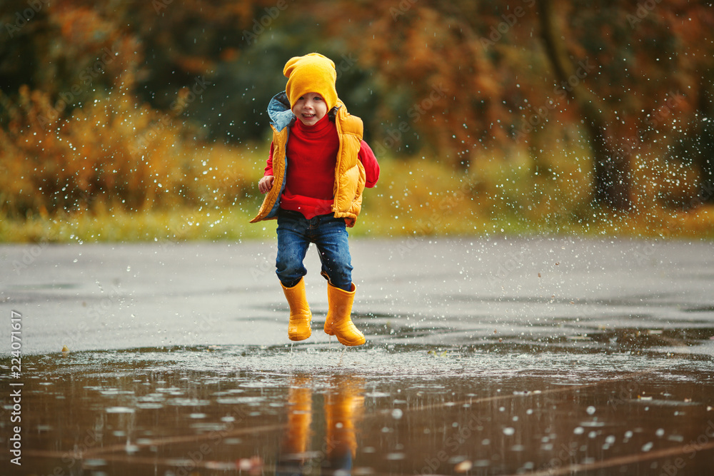 穿着橡胶靴的快乐小男孩在秋天散步时跳到水坑里