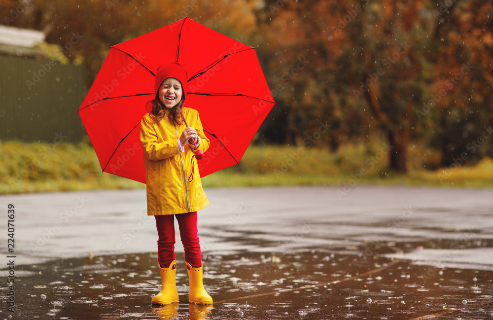 带着雨伞和橡胶靴的快乐小女孩在秋天散步时跳到水坑里