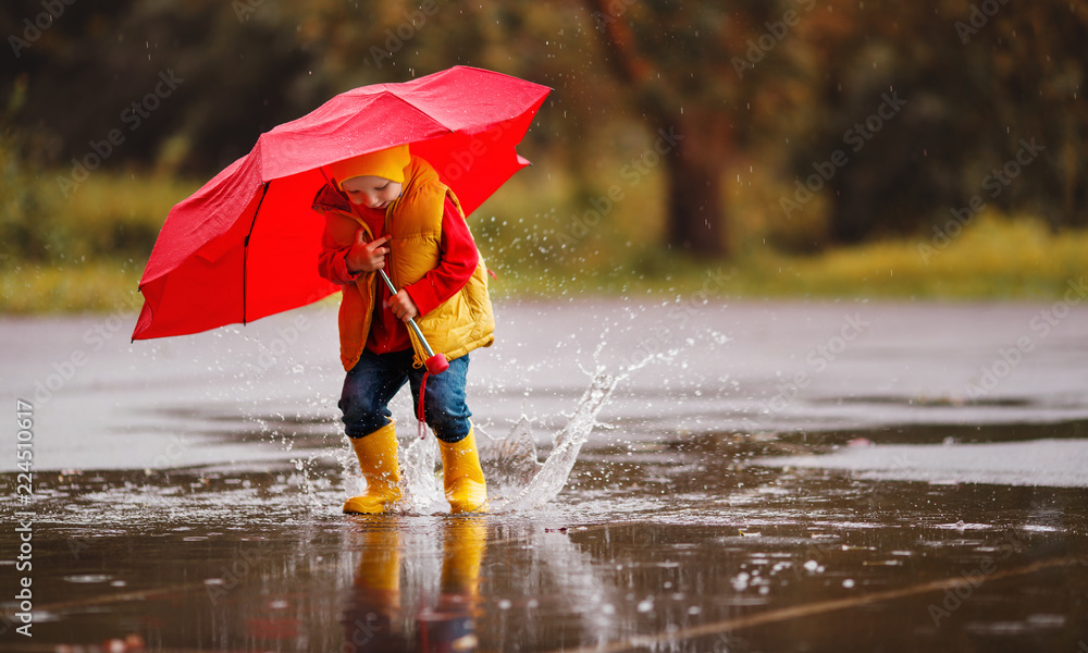 快乐的小男孩，穿着橡胶靴，打着雨伞，在水坑里跳秋天的散步