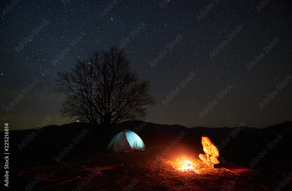 山中露营之夜。美丽的星空下，年轻的女游客在燃烧的篝火旁休息