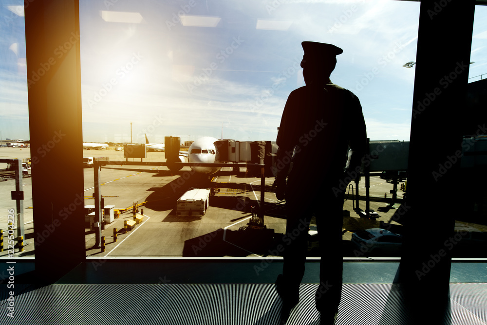身穿制服的飞行员站在机场航站楼登机口区域，正望向窗外。