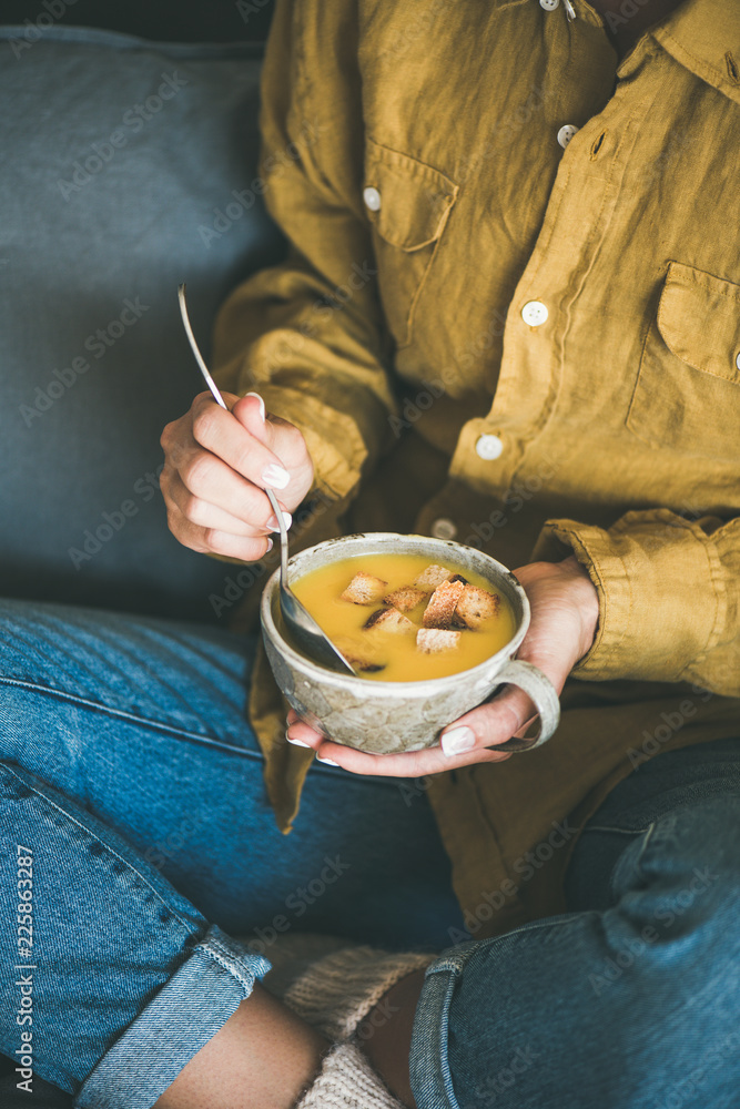 穿着牛仔牛仔裤和芥末衬衫的女性坐着，端着一杯温暖秋天的南瓜奶油汤