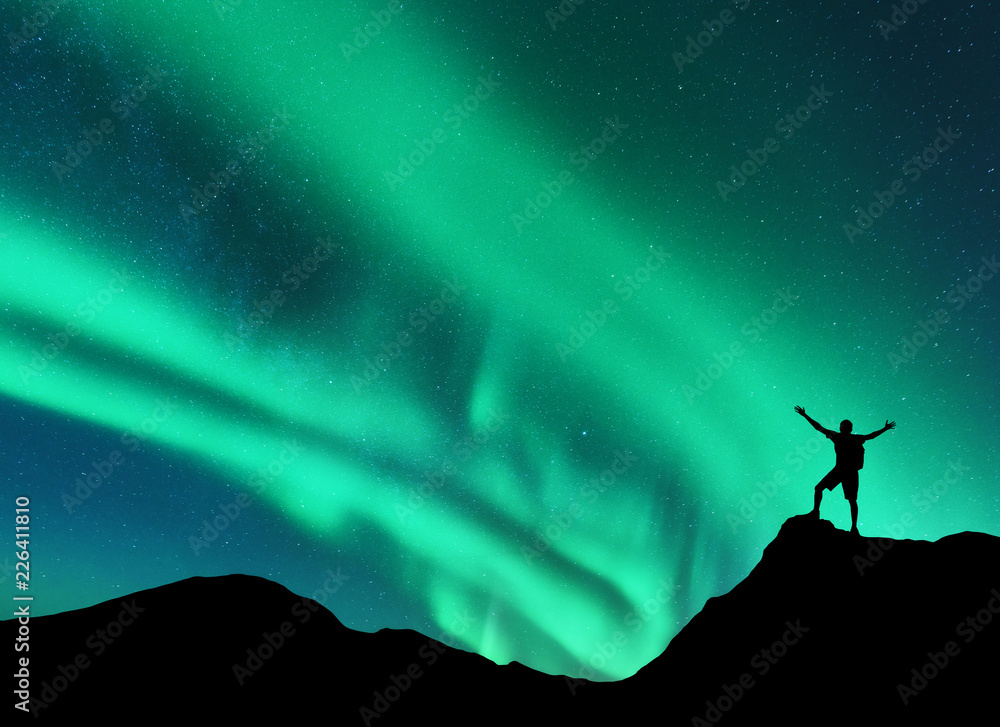 挪威山峰上举起双臂站立的人的北极光和剪影。A