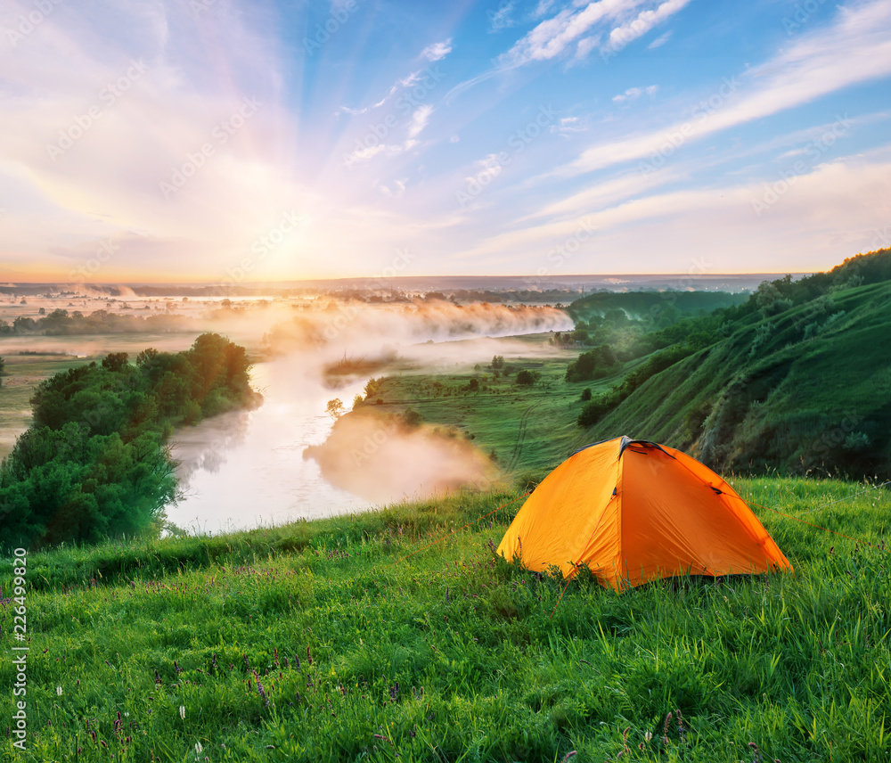 河流上方绿草如茵的山丘上的橙色旅游帐篷