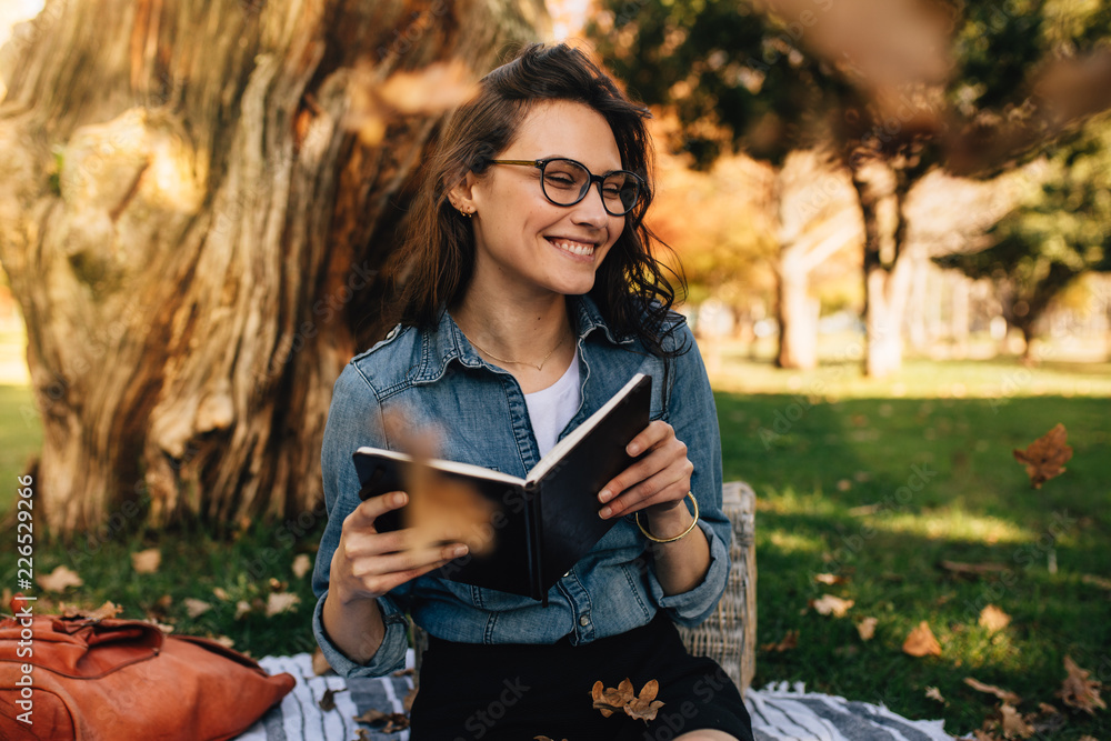 微笑的女人坐在公园里，手里拿着书，树叶飘落