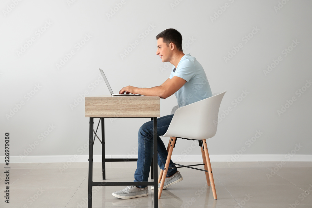 年轻人坐在靠着浅色墙壁的桌子旁，拿着笔记本电脑工作