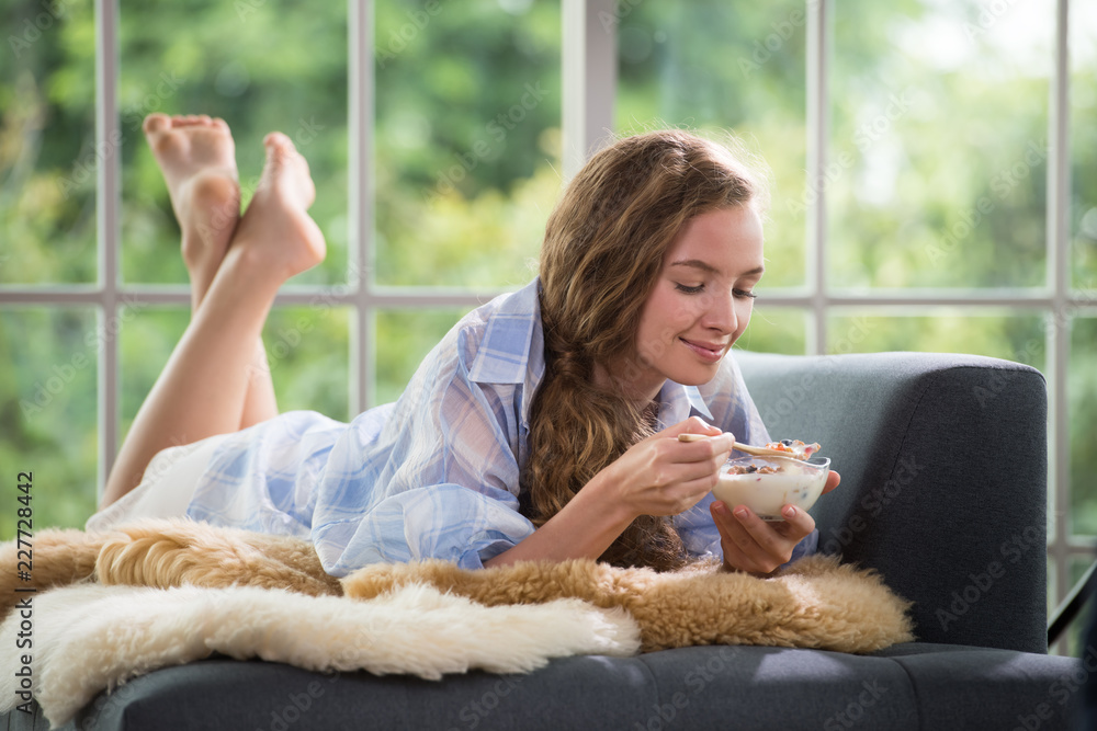 健康的年轻女人躺在沙发上，手里拿着一碗酸奶，看起来轻松舒适