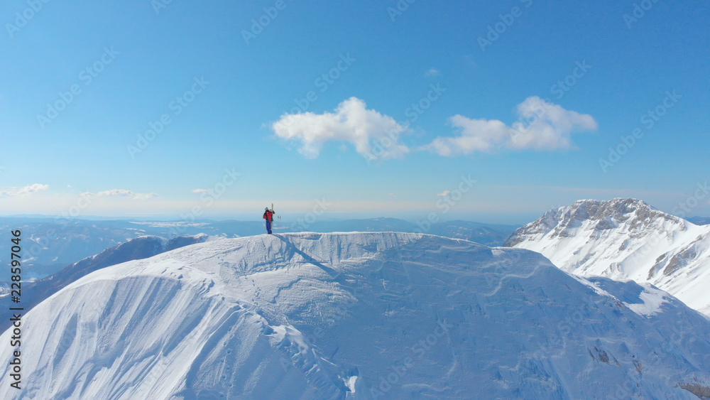 AERIAL: Unrecognizable skier taking photos after reaching peak of snowy mountain