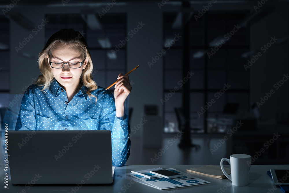 在黑暗的办公室里，漂亮的金发女郎在笔记本电脑上工作，并得到一个