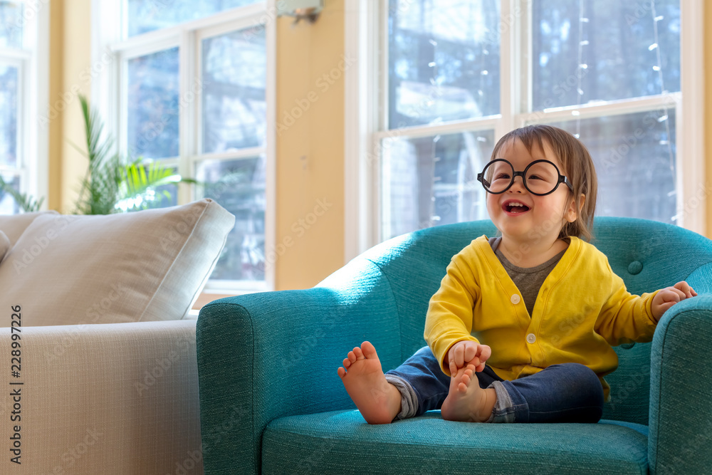 快乐的蹒跚学步的男孩戴着眼镜在大椅子上玩耍