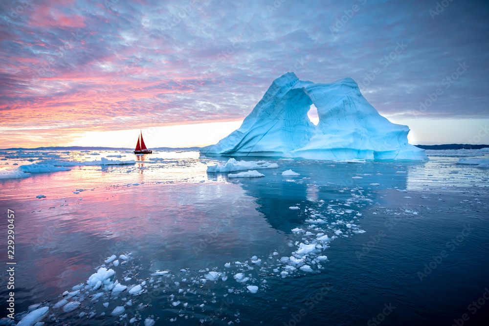 Little red sailboat cruising among floating icebergs in Disko Bay glacier during midnight sun season