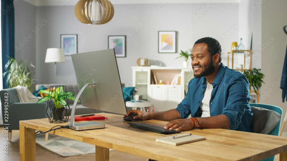 英俊的黑人坐在舒适客厅的桌子旁用个人电脑工作。