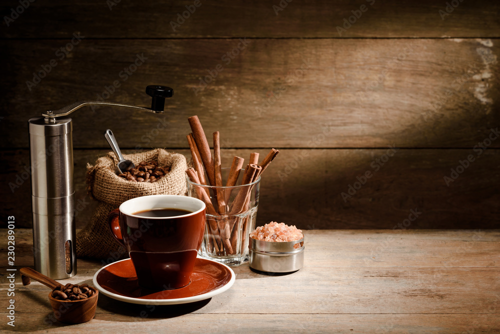 A Cup of black coffee with coffee bean bag, cinnamon, coffee grinder and sugar on wooden floor with 