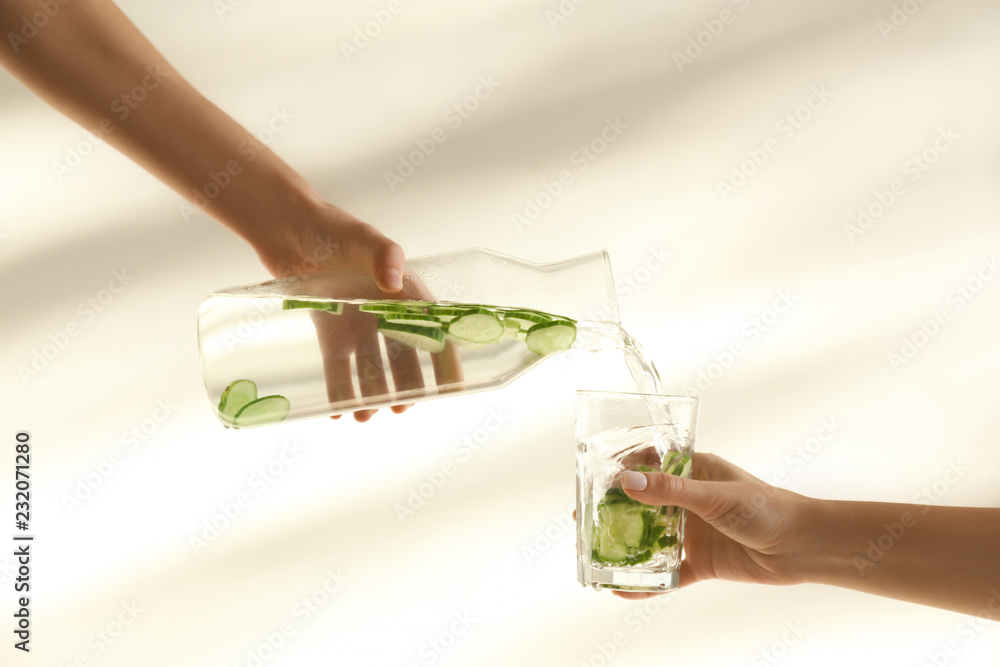 一名女子在浅色背景下将新鲜的黄瓜水从瓶子里倒进玻璃杯