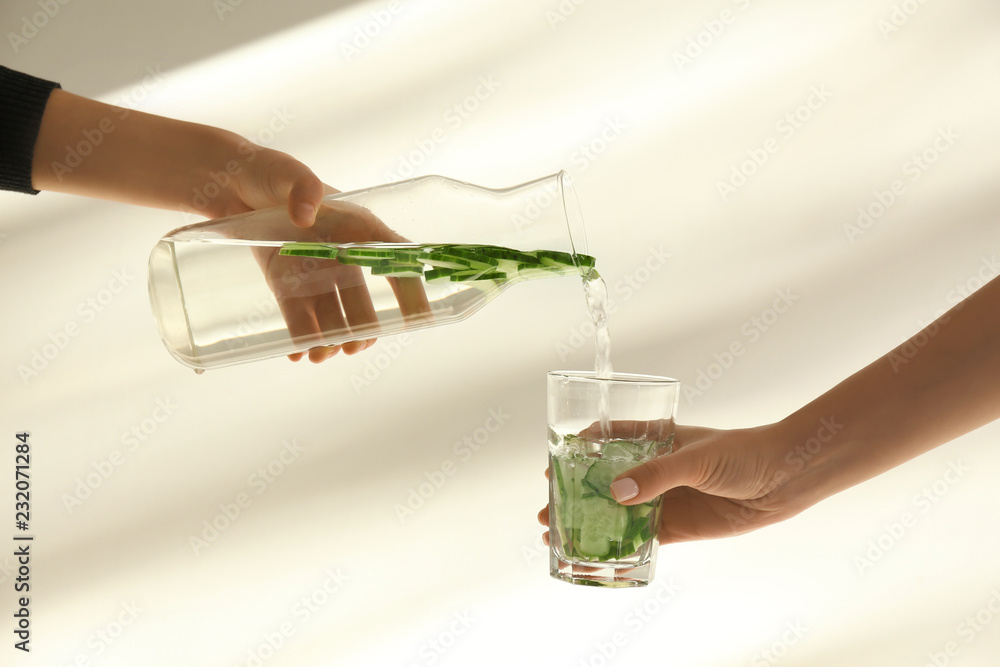 女子在浅色背景下将新鲜的黄瓜水从瓶子里倒进玻璃杯