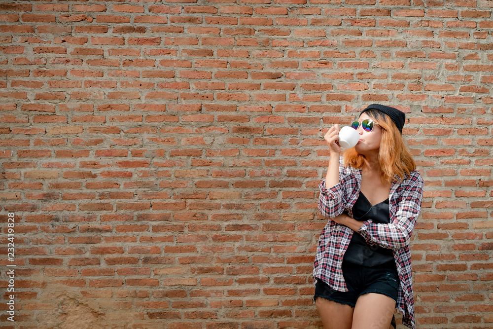 快乐的年轻潮人女人站在镇街的砖墙旁，手里拿着咖啡杯。