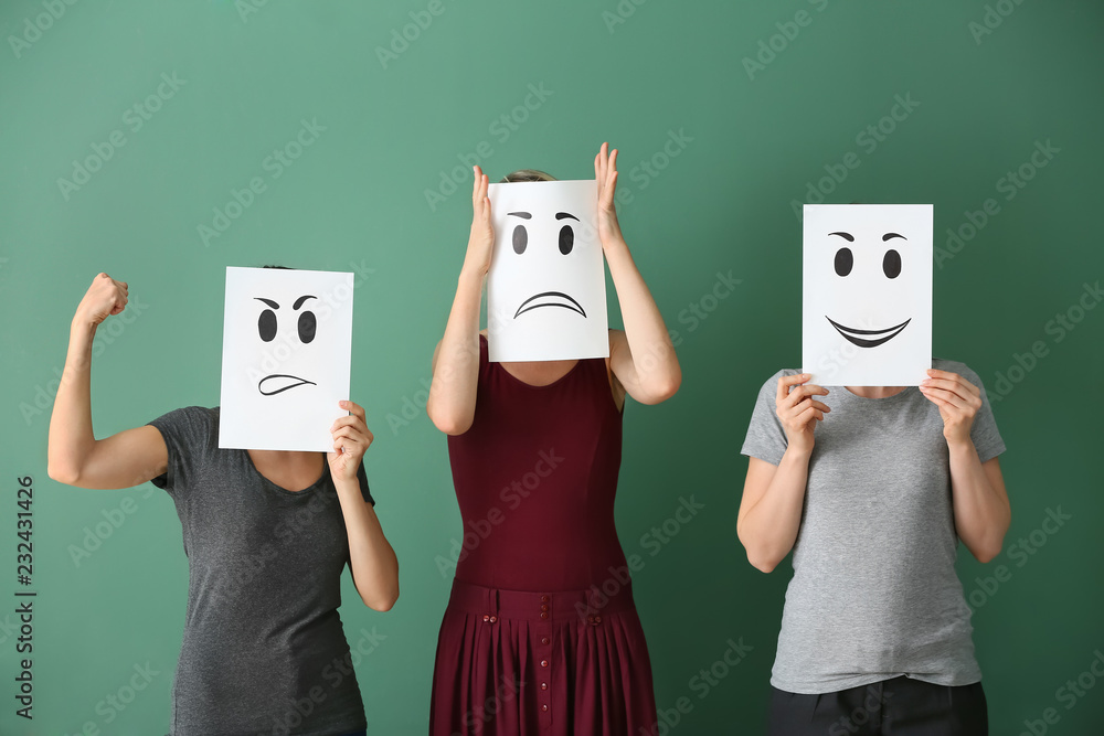 Young women hiding faces behind sheets of paper with drawn emoticons on color background