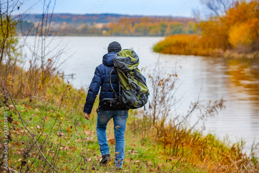 A tourist with a backpack walks next to the river in autumn. A beautiful landskape of nature in yell
