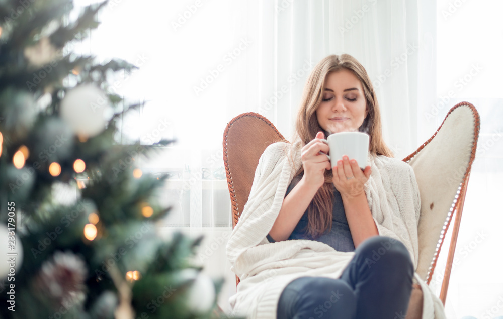 年轻放松的女人在圣诞树旁休息，喝一杯热茶或咖啡