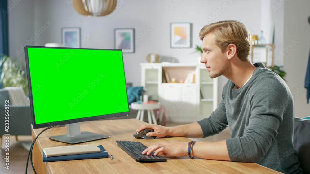 英俊的年轻人坐在办公桌前，在绿色实体屏幕个人电脑上工作
